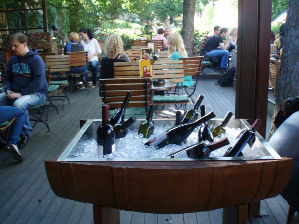 Gosti puba na zunanji terasi za lesenimi mizami. V ospredju hladilna posoda z vini, narejena iz soda.