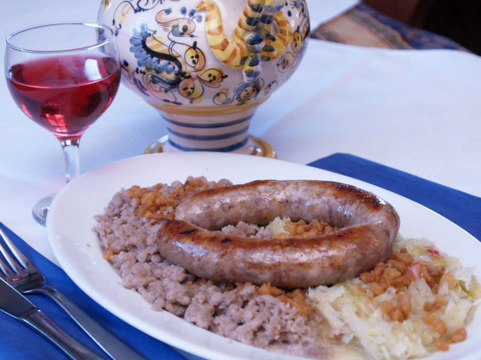 Pečenica, s kislim zeljem in ajdovo kašo. V ozadju majolka in kozarec vina. 