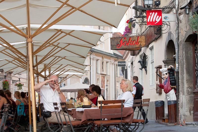 Gostje sedijo za mizami pod senčniki na terasi pred lokalom.