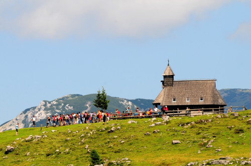 Skupina ljudi pred cerkvijo. V ospredju travnik. V ozadju planine. 