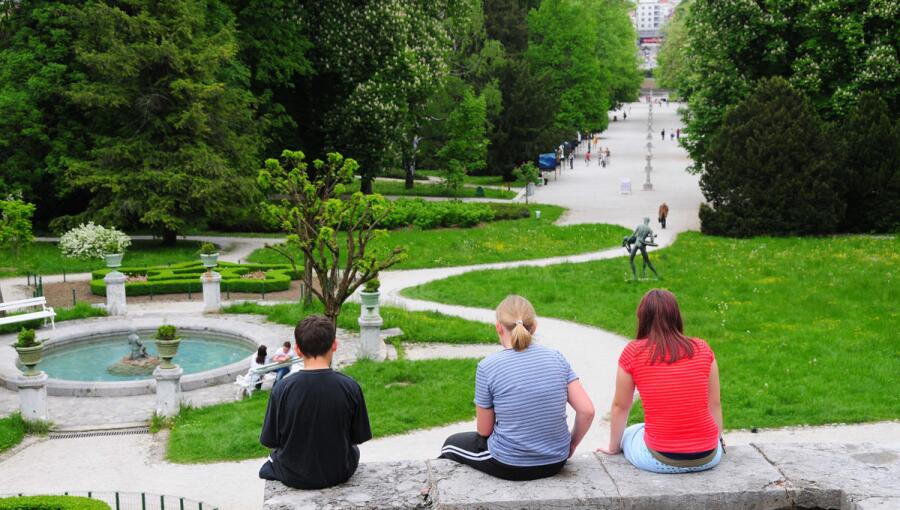 Three young people are sitting on a stone wall, observing the walking paths, a fountain, and the neatly landscaped park in the background.