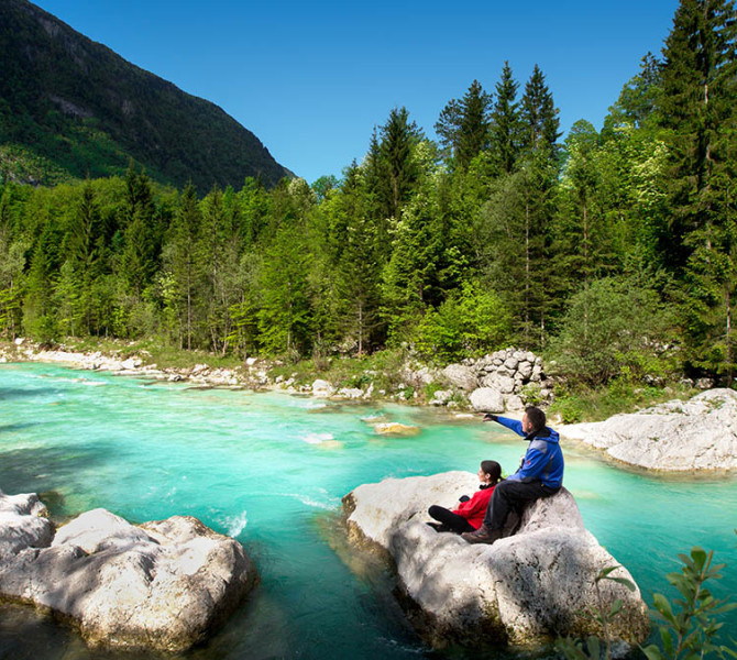 Smaragdna reka obdana z gozdom. Na veliki skali sedita dva obiskovalca. 