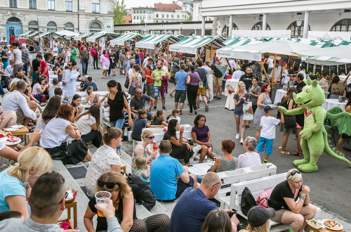 Maskota Ljubljanski zmajček na Odprti kuhni. Ljudje sedijo na klopicah in jedo hrano.