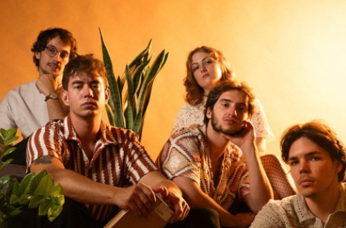 The band is sitting on a sofa in front of an orange background, posing for a photo.