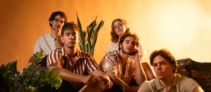 The band is sitting on a sofa in front of an orange background, posing for a photo.