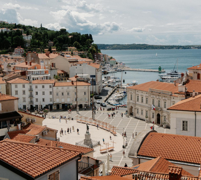 Luftaufnahme des Tartini-Platzes in Piran mit roten Ziegeldächern, Fußgängern und dem Hafen am Meer im Hintergrund.