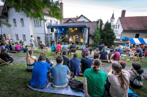 Ljudje sedijo na travi ter gledajo in poslusajo koncert.