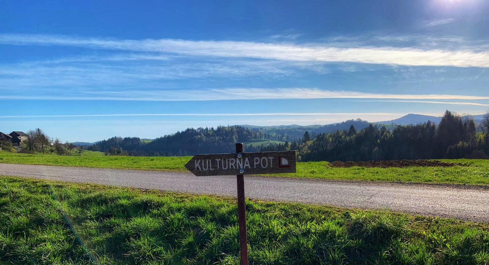 The path winds past green meadows, surrounded by hills, with the blue sky stretching above.