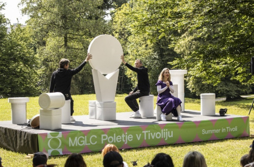 Predstava na odru, postavljenem na travniku, kjer igralci nastopajo z razlicnimi belimi kipci.