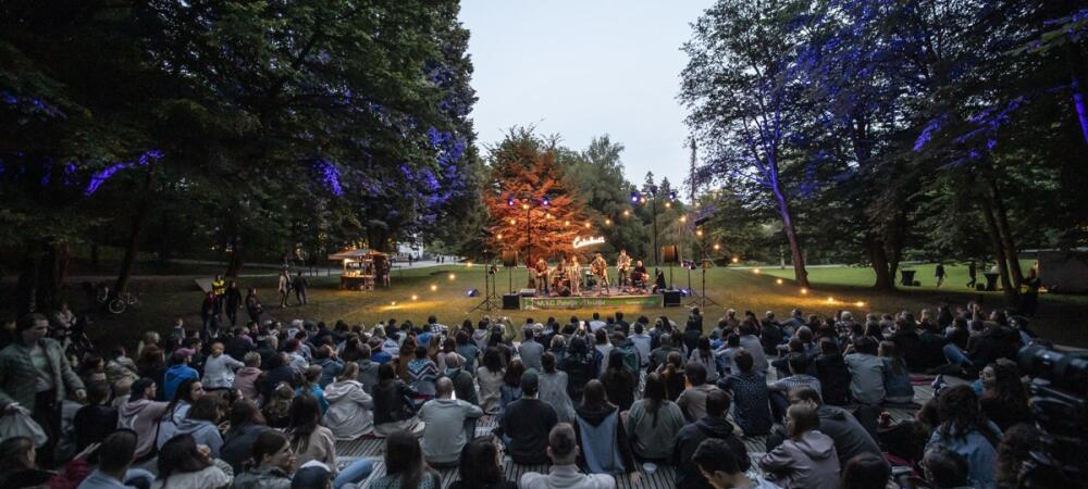 A large audience gathers in a park at dusk to watch a live band perform on an outdoor stage, illuminated by decorative lights among the trees.