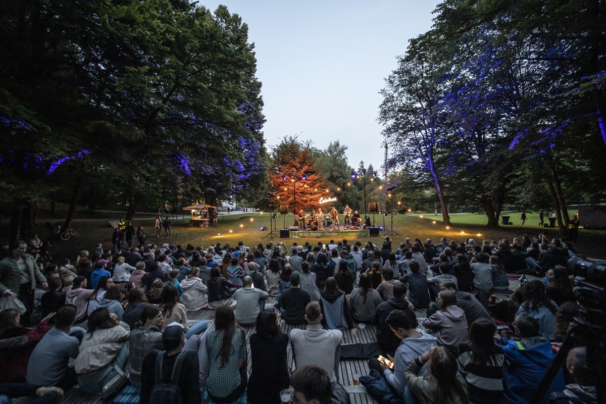 A large audience gathers in a park at dusk to watch a live band perform on an outdoor stage, illuminated by decorative lights among the trees.