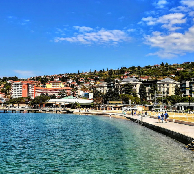 A promenade with people by the calm blue sea in Portoroz, with hotels and houses on a green hillside in the background.