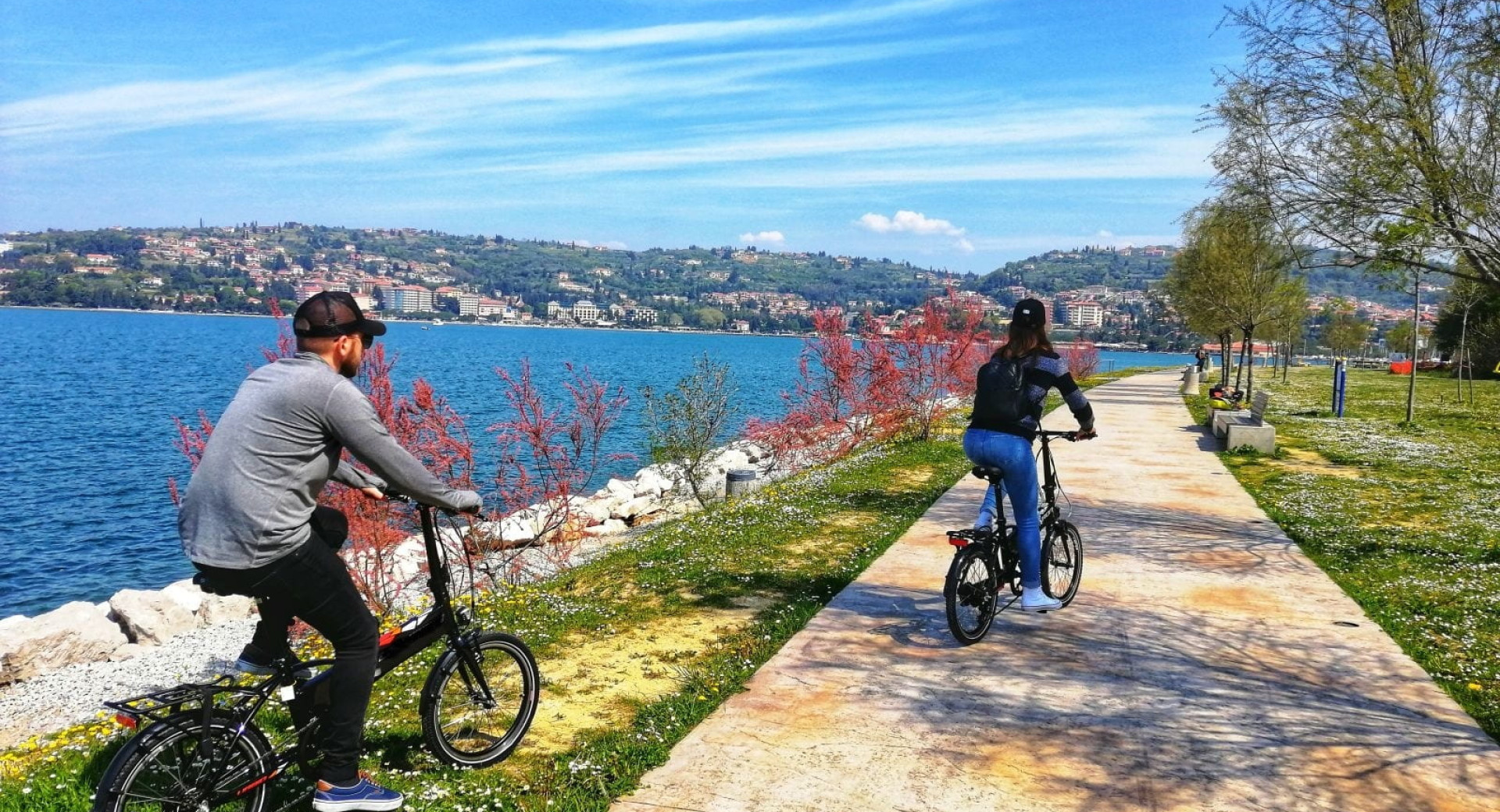 Two cyclists ride along a coastal promenade in Lucija, with the sea and Portoroz in the background.
