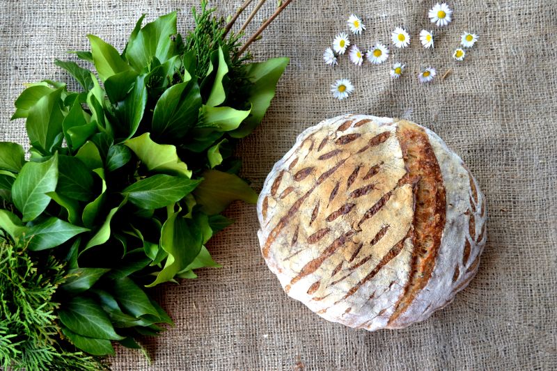On the table with a linen cloth, there is a loaf of bread, daisy flowers, and green leaves in a bouquet.