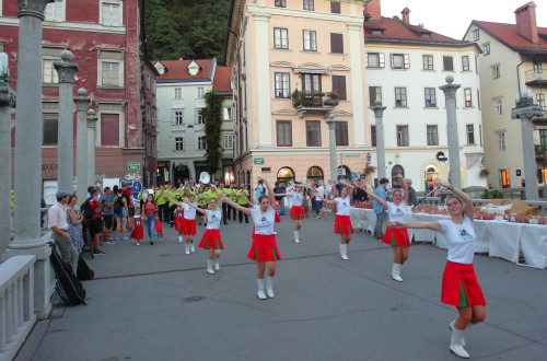 A parade of a marching band and majorettes winds its way through the city, crossing a bridge over the Ljubljanica River, with Ljubljana Castle rising in the background.