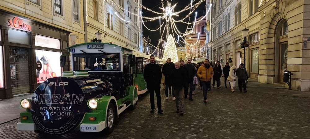 A city train in black and green stands in a festive town decorated with lights.