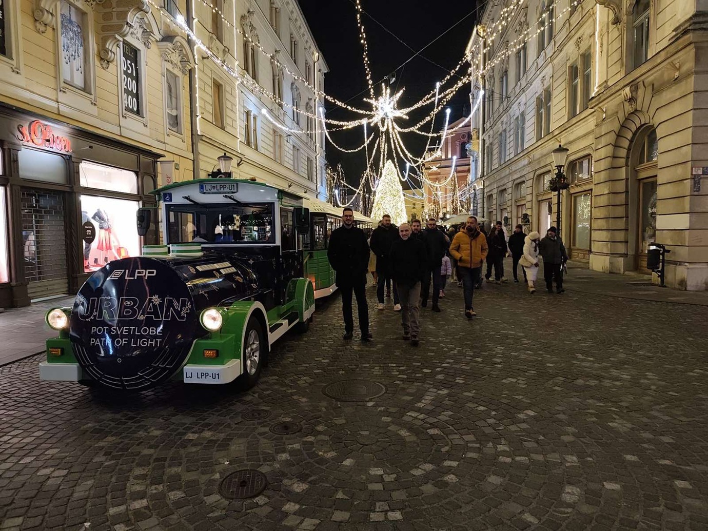 Un train urbain noir et vert se tient dans une ville décorée de lumières pour les fêtes.