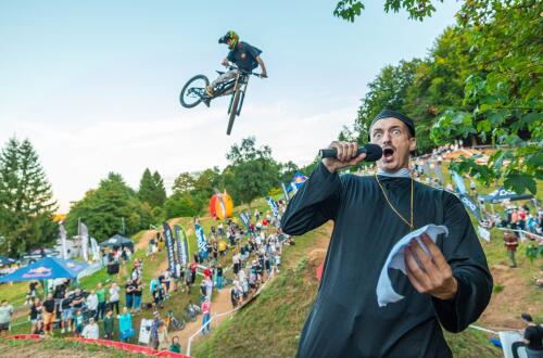 A man in a black robe in the foreground speaks into a microphone while a cyclist in the background performs a jump at an outdoor event surrounded by trees and spectators.