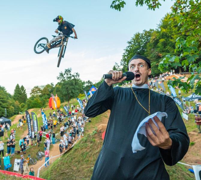A man in a black robe in the foreground speaks into a microphone while a cyclist in the background performs a jump at an outdoor event surrounded by trees and spectators.