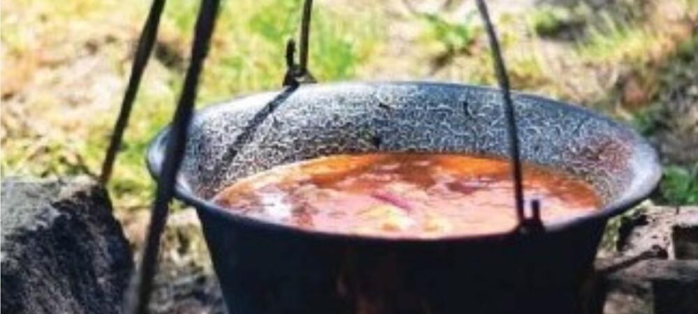 Črn lonec visi nad odprtim ognjem, poln golaža, v ozadju pa je mogoče videti travo in zemljo, kar nakazuje kuhanje na prostem.