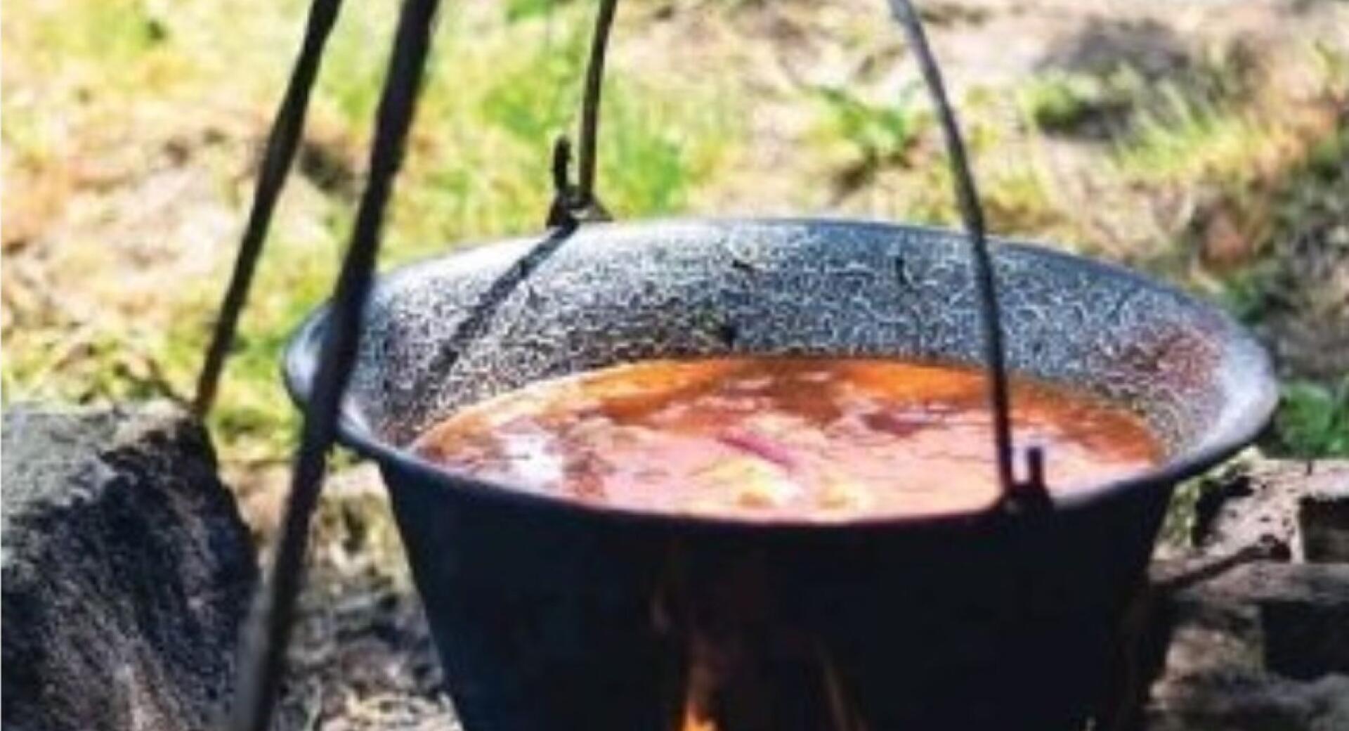Črn lonec visi nad odprtim ognjem, poln golaža, v ozadju pa je mogoče videti travo in zemljo, kar nakazuje kuhanje na prostem.