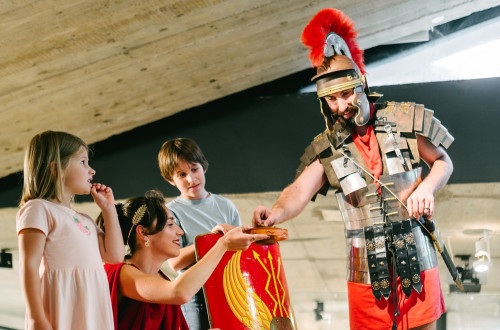 Two adults dressed as ancient Romans and two children are in a room with sand and gravel on the floor. The boy holds a knight’s shield, and the man is passing something to the woman on a wooden tray.