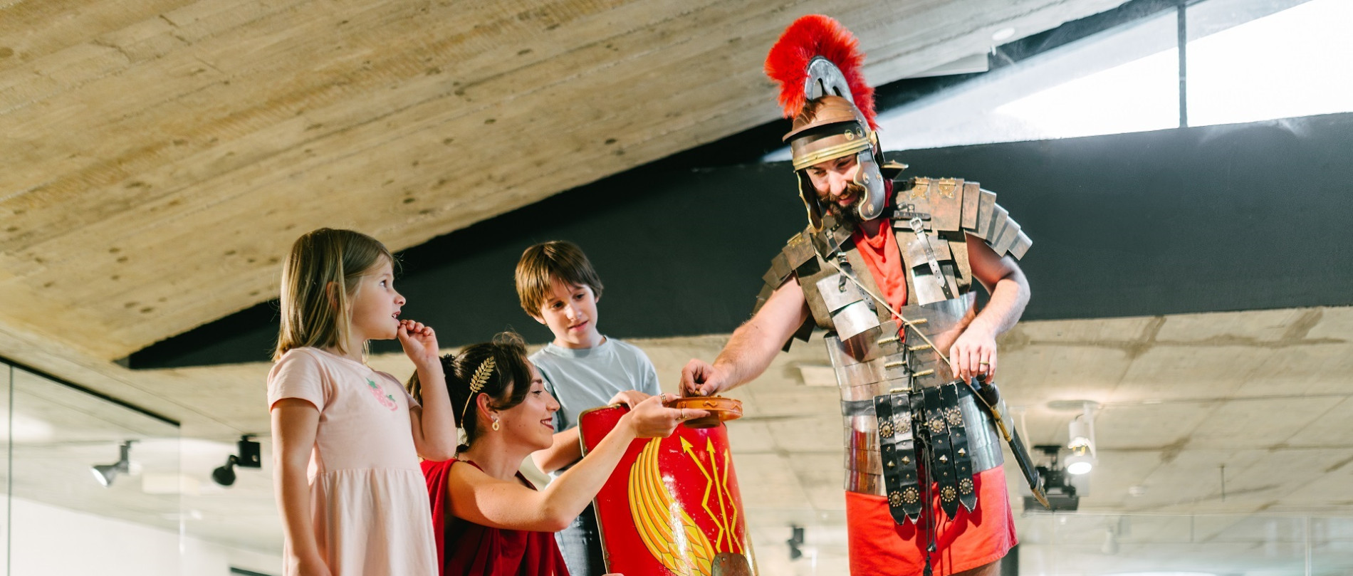 Two adults dressed as ancient Romans and two children are in a room with sand and gravel on the floor. The boy holds a knight’s shield, and the man is passing something to the woman on a wooden tray.