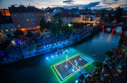 V mraku se s pticje perspektive razprostira pogled na Ljubljanico, na kateri je postavljeno odbojkarsko igrisce, rahlo potopljeno v vodo, in osvetljeno z raznobarvnimi lucmi.