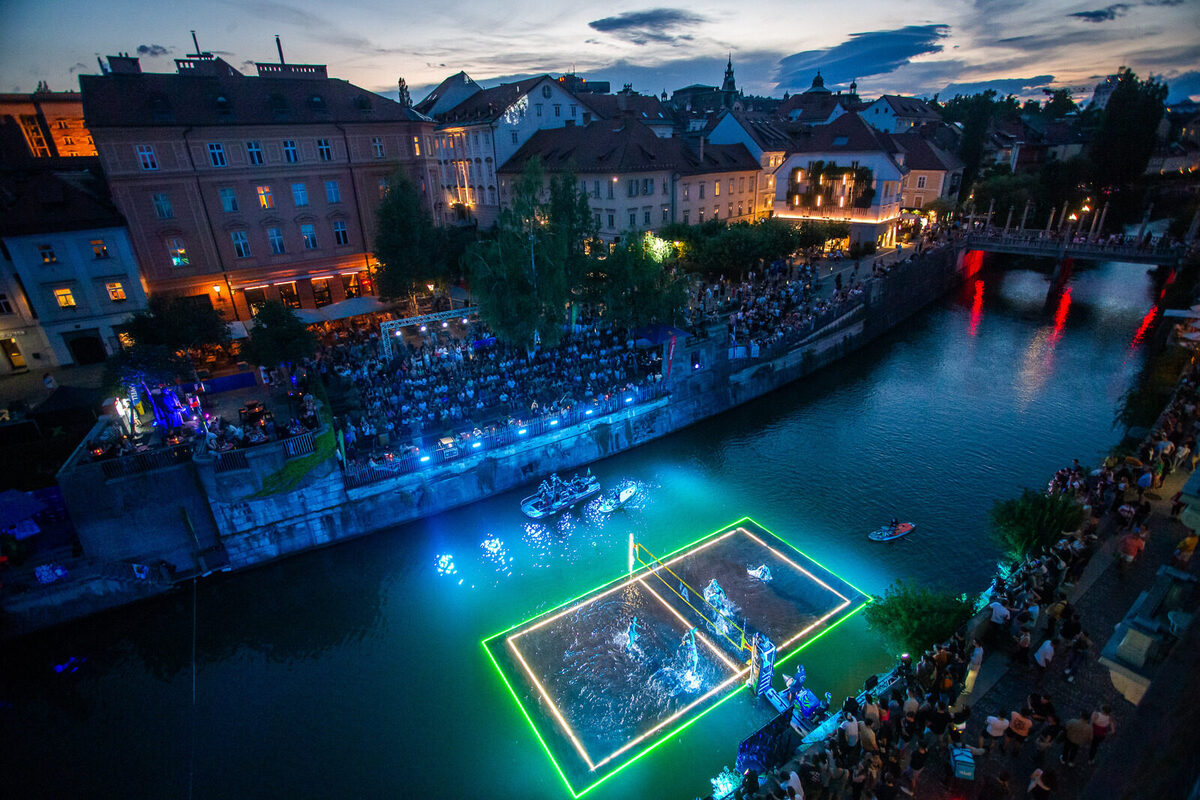 V mraku se s pticje perspektive razprostira pogled na Ljubljanico, na kateri je postavljeno odbojkarsko igrisce, rahlo potopljeno v vodo, in osvetljeno z raznobarvnimi lucmi.