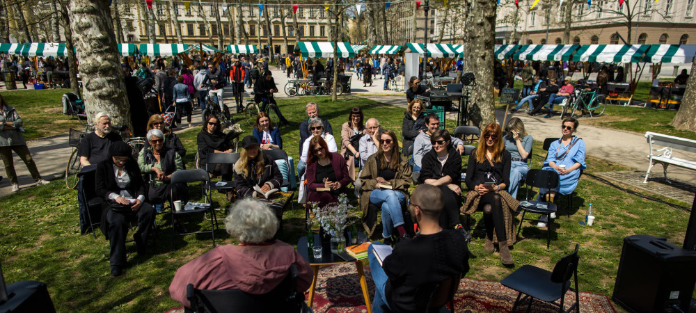 People are sitting in the park, with wooden stalls featuring white and green roofs in the background.