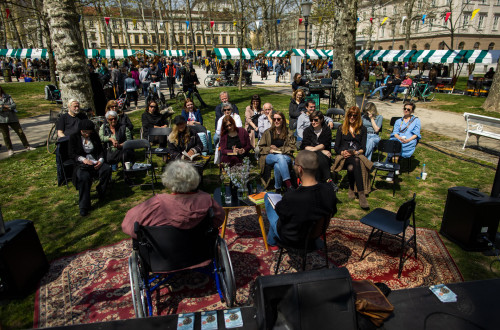 People are sitting in the park, with wooden stalls featuring white and green roofs in the background.