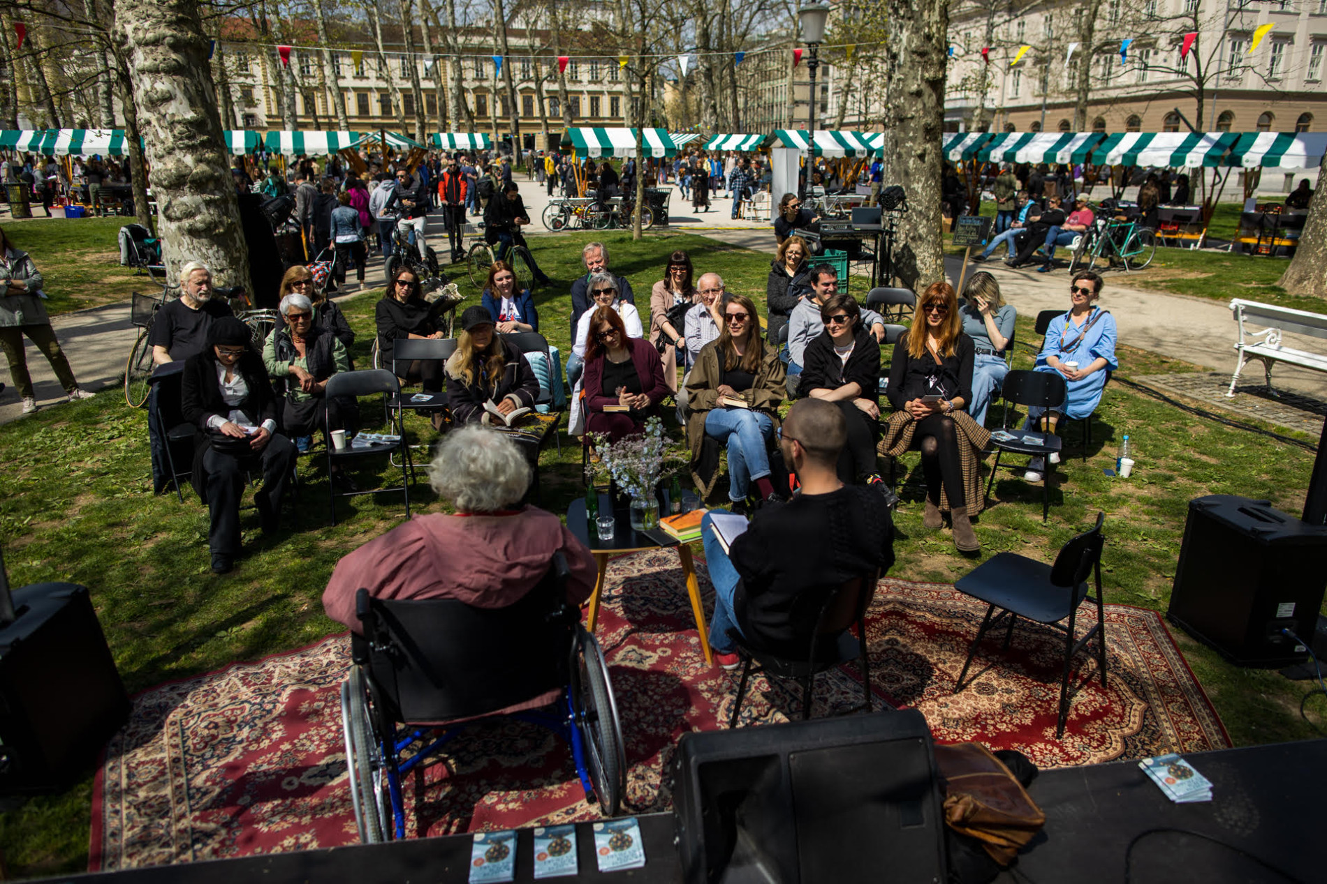 People are sitting in the park, with wooden stalls featuring white and green roofs in the background.