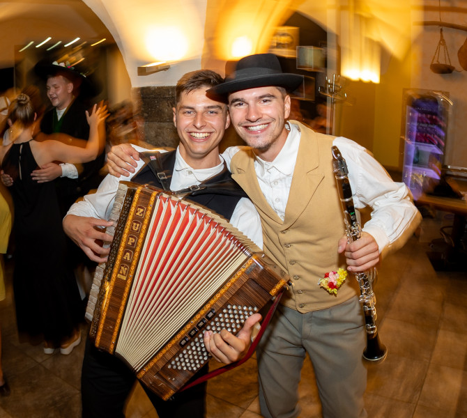 Two male musicians smile as they pose for a photo — one is playing the accordion, and the other is holding a clarinet.