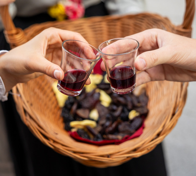 A woman’s hand and a man’s hand clink glasses filled with drinks; in the background, there is a blurred basket of dried fruit.