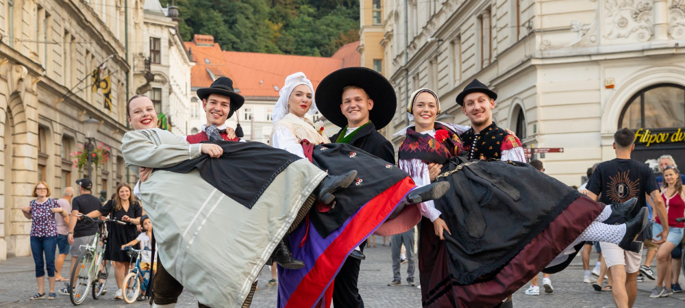 Trije pari v narodnih nosah so sredi mesta, kjer moski drzijo v narocju tri zenske.