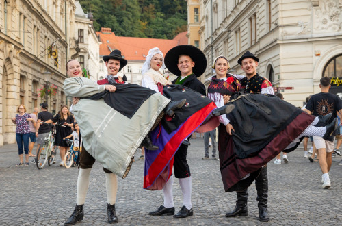 Three couples in traditional costumes are in the city centre, where the men are holding three women in their arms.