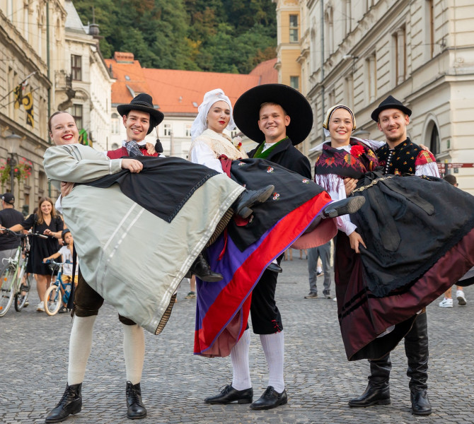 Three couples in traditional costumes are in the city centre, where the men are holding three women in their arms.