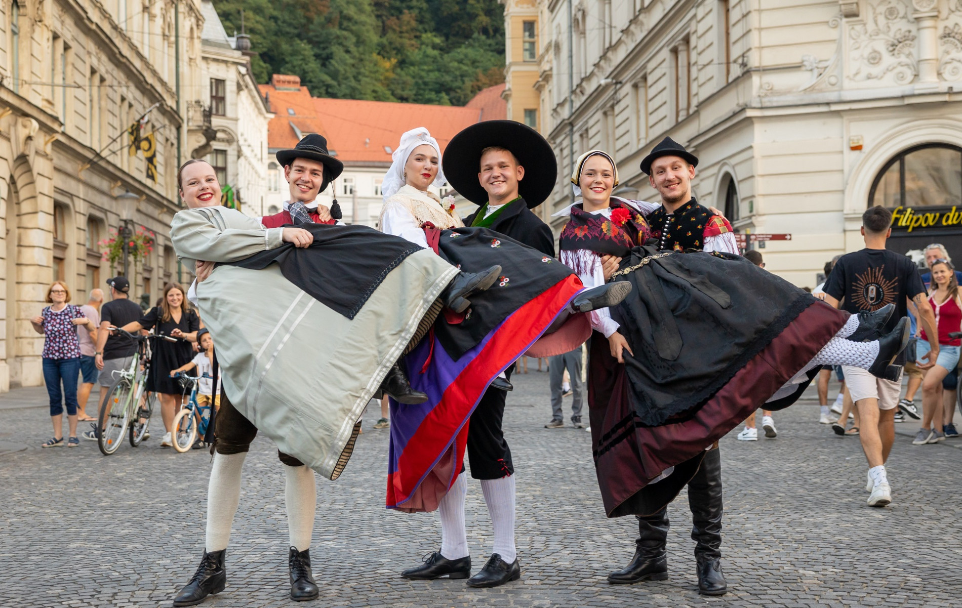Three couples in traditional costumes are in the city centre, where the men are holding three women in their arms.