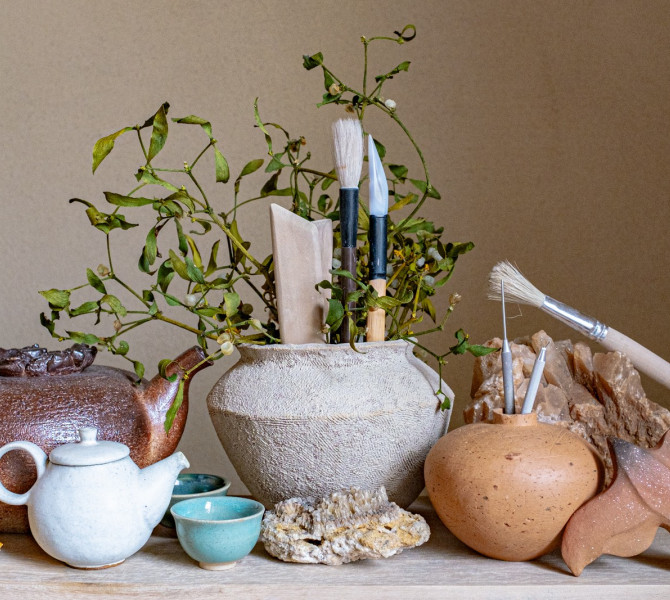 The scene shows a composition of handmade ceramics and a vase filled with sculpting tools and green leaves.