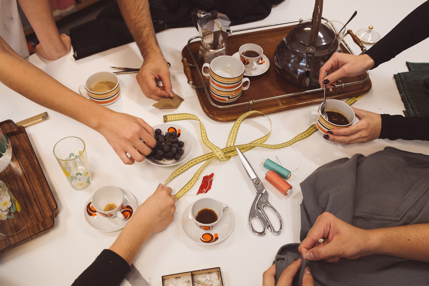 A work table where different hands work together to create something. On the table there are tea cups, measuring tape, scissors and thread.