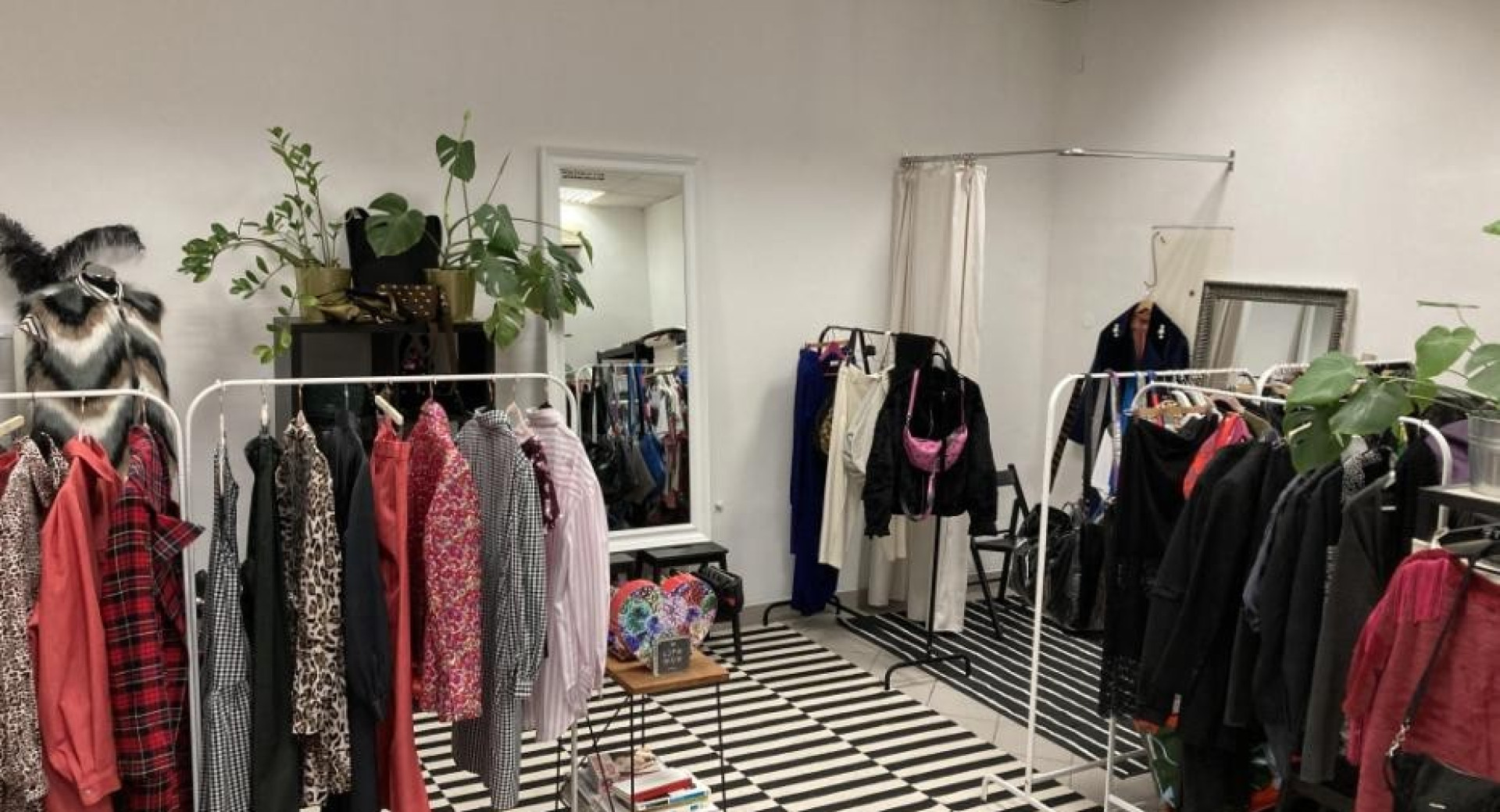 The boutique displays colourful clothes on white racks.  A mirror, a small table with magazines and a curtained fitting area complete the space.