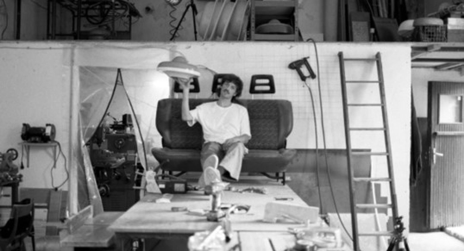 Na kavcu v delavnici sedi oseba, ki v eni roki drzi svetilko. Prostor je zapolnjen z razlicnim orodjem in opremo.