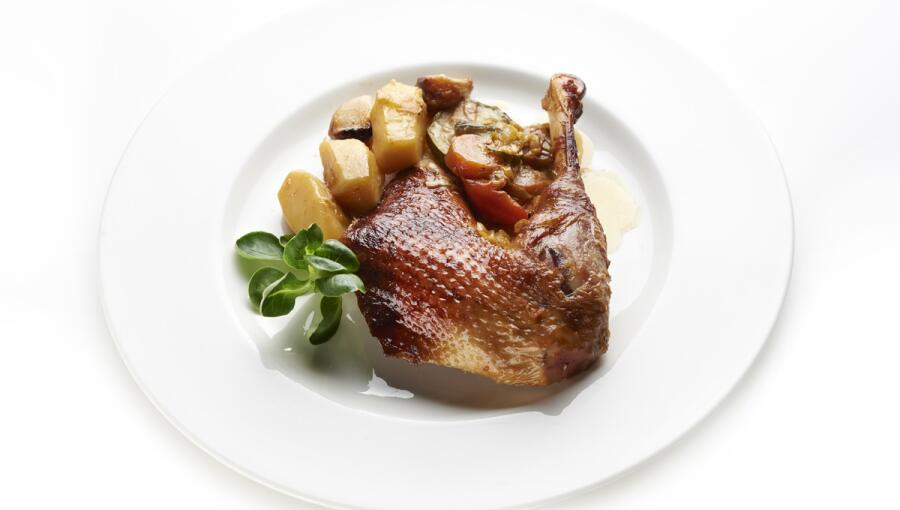 On a white plate, there is a nicely roasted duck leg accompanied by a variety of roasted vegetables, including potatoes and carrots, all garnished with fresh green herbs.