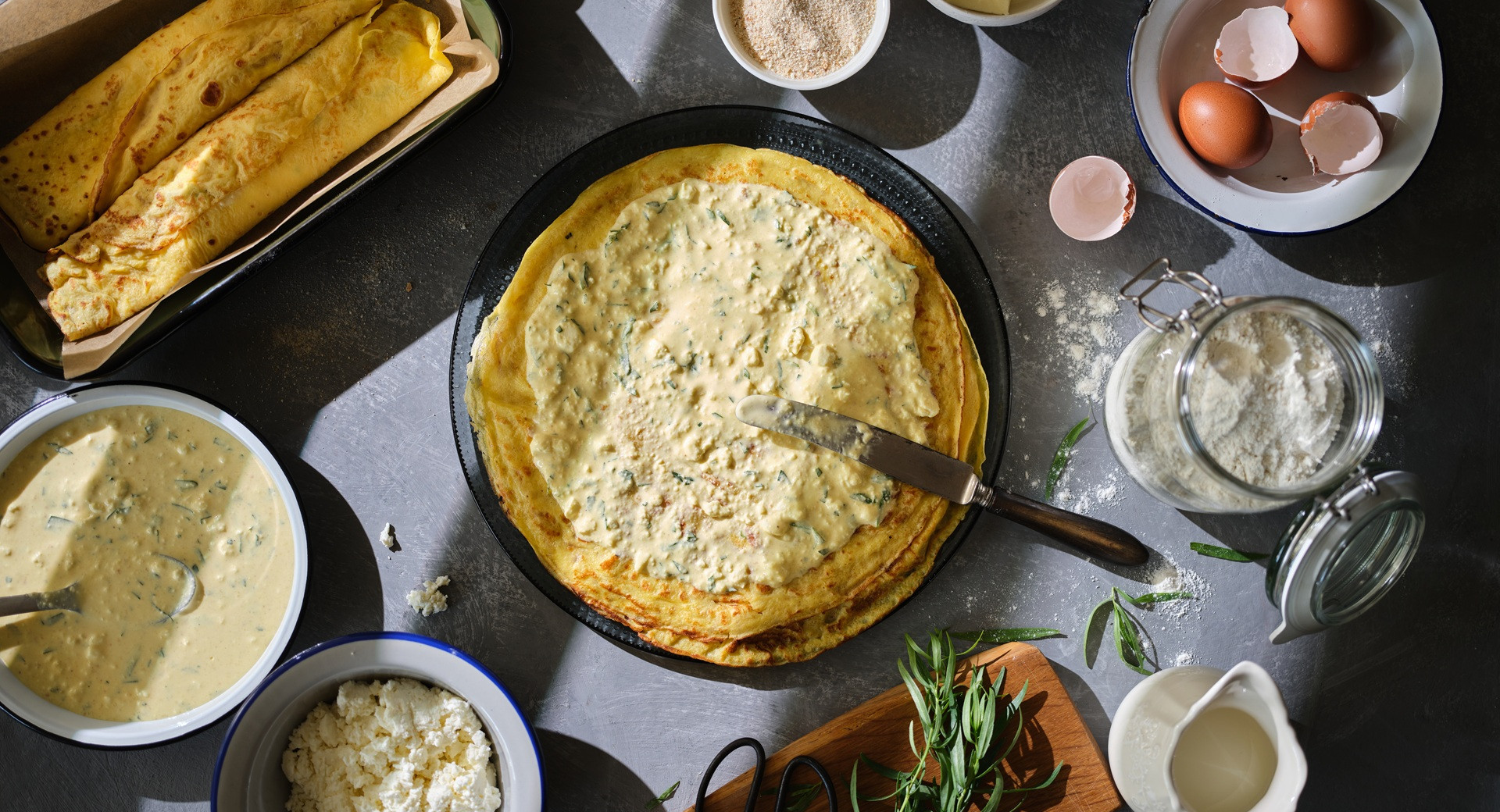 A tarragon pancake spread with cottage cheese on a black plate. Ingredients for the pancakes in small bowls.
