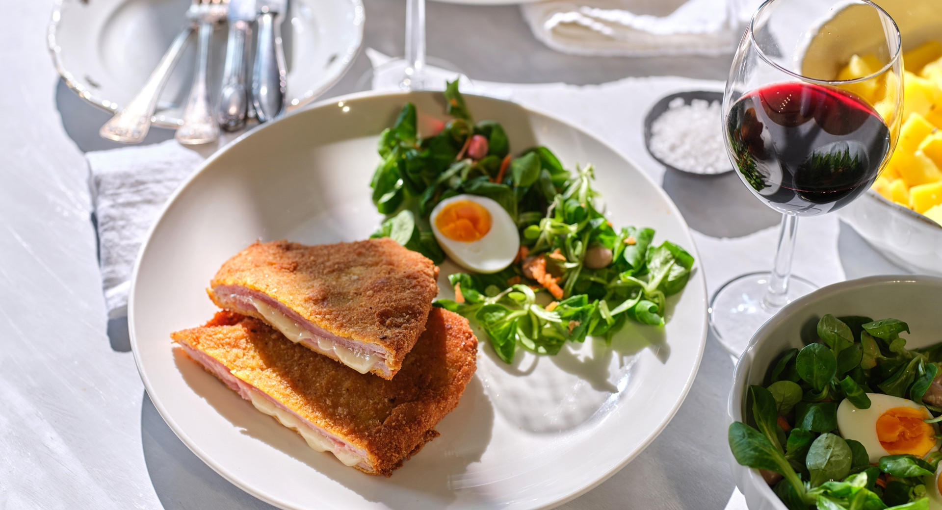 On the plate are two pieces of crispy fried schnitzel with ham and cheese, surrounded by fresh salad and a halved boiled egg, accompanied by a glass of red wine.