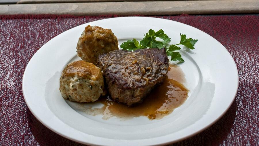 A plate featuring a portion of beef, accompanied by two dumplings and garnished with fresh parsley. There is a rich gravy surrounding the meat.