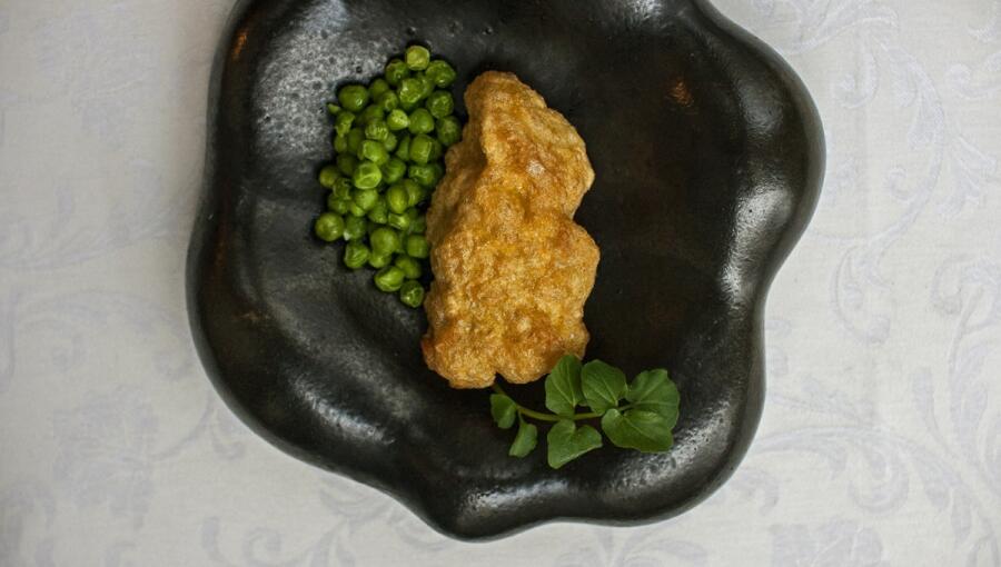 Golden fried food on a black plate is accompanied by fresh green peas and a sprig of herbs.