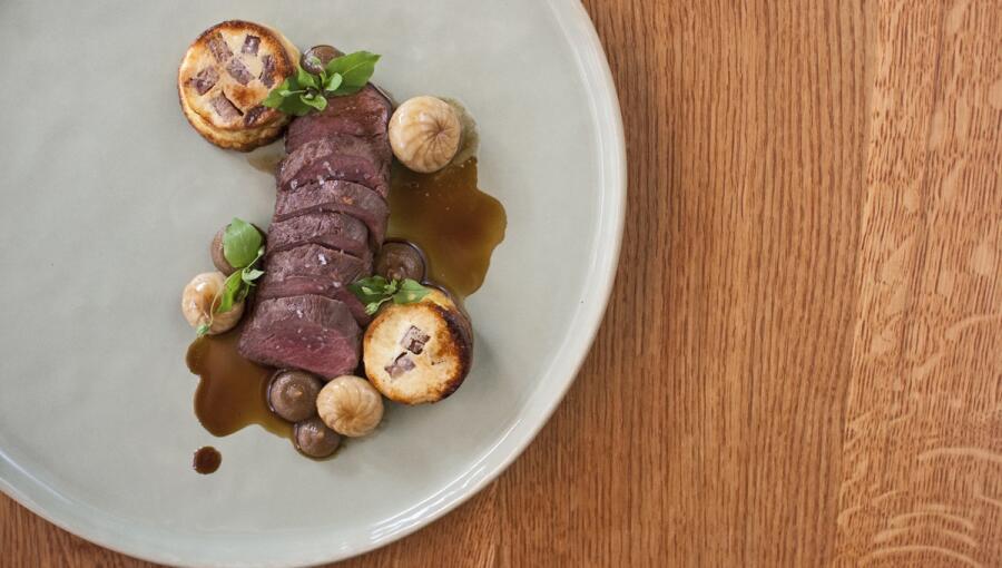 On the plate, there are slices of juicy meat, surrounded by potatoes and small mushrooms, garnished with herbs and drizzled with sauce.