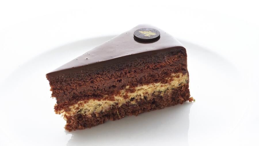 A slice of dark chocolate cake with moist chocolate layers and a creamy filling, topped with a shiny chocolate glaze and decorative chocolate piece.