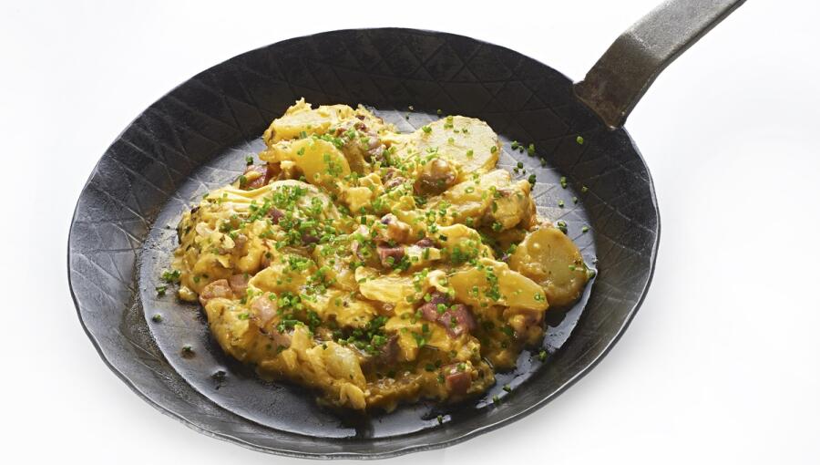 In the pan is a dish that includes potatoes, pieces of meat, and a sprinkle of fresh chives, set against a plain white background.